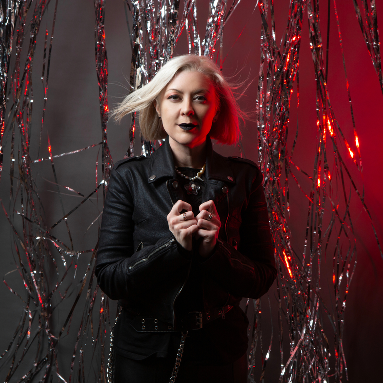 Cover of 'The Longest Time' EP. Laura is standing facing the wind, her blonde hair is flowing out behind her and she is holding the collar of her black biker jacket. There is silver foil strips blowing in the air behind her and the backdrop is red and black.